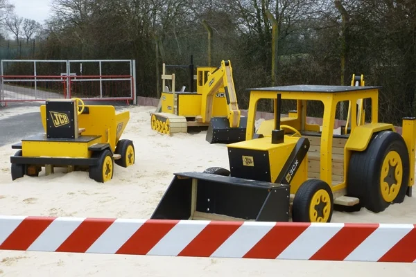A yellow and black timber play JCB digger in a construction-themed sand pit at an outdoor playground.