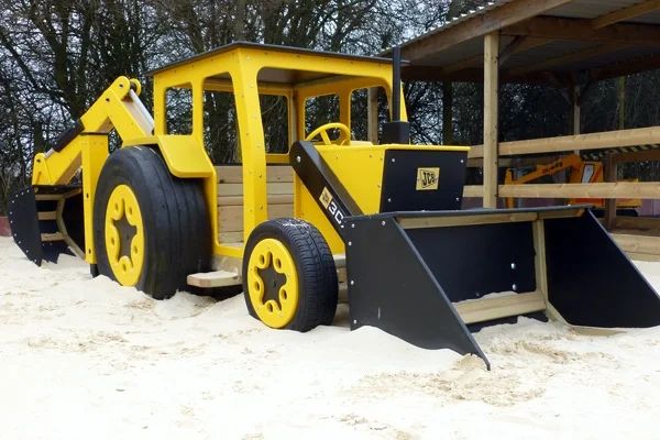 A yellow and black timber play JCB digger with a front bucket and rear digging arm, in a sand pit at an outdoor playground.