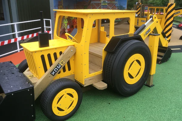 A side view of a yellow and black timber play JCB digger on a rubber surface in an outdoor playground.