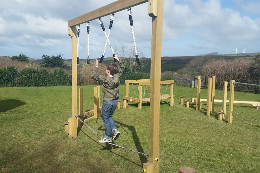 A child reaching up to grip the hanging ropes on a Drop Rope Traverse outdoor play equipment, with a trim trail course visible in the background.