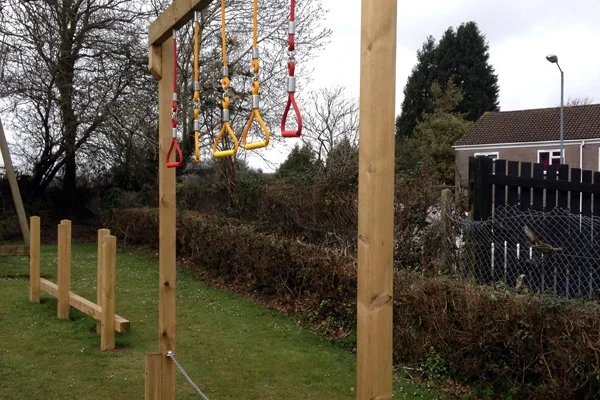 A close-up of a Drop Rope Traverse showing colourful red and yellow triangular ring handles hanging from a wooden frame in a garden setting.