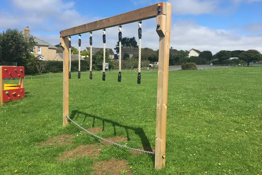 A wooden Drop Rope Traverse outdoor play equipment with hanging ropes and coloured bolt caps, installed in a sunny school field.