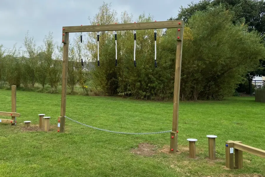 A Drop Rope Traverse with hanging ropes installed on a green grass playground, with stepping posts and other trim trail equipment visible nearby.