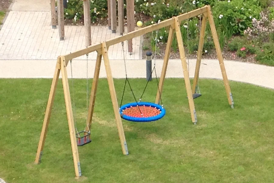 An aerial view of a timber triple swing frame with a blue and red nest swing, a red toddler seat, and a black flat seat, set on green grass in an outdoor playground.
