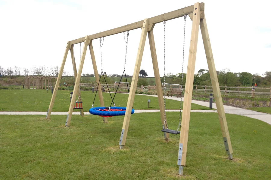 A timber triple swing frame with a blue and red nest swing, a red toddler seat, and a black flat seat, set on green grass in an outdoor playground.