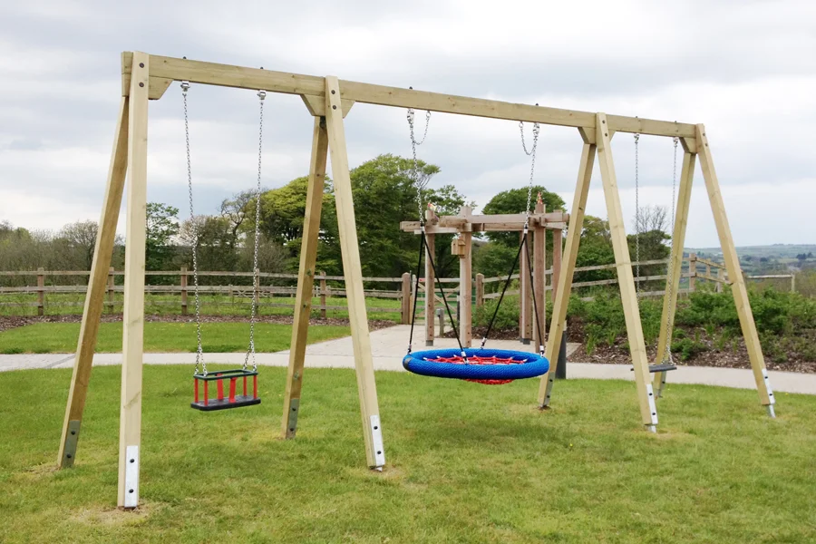 A wide view of a timber triple swing frame with a blue and red nest swing, a red toddler seat, and a black flat seat, set on green grass in an outdoor playground with trees and a wooden fence.