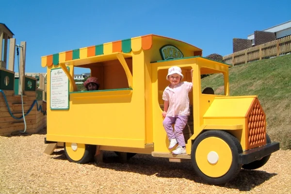 A yellow timber play ice cream van with a green and orange striped awning, with a girl standing in the doorway, in a woodchip outdoor playground.
