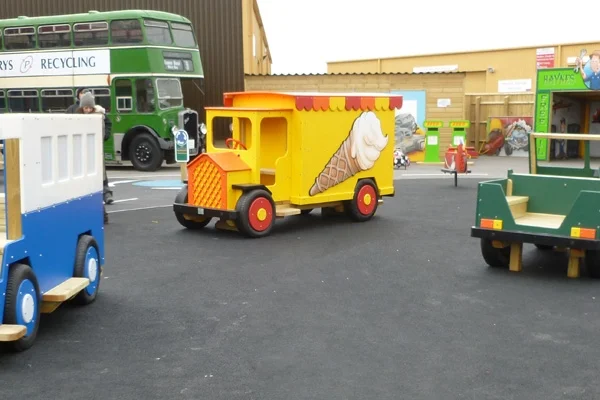 A front view of a yellow and orange timber play ice cream van with an ice cream cone artwork, on a tarmac surface in an outdoor playground.