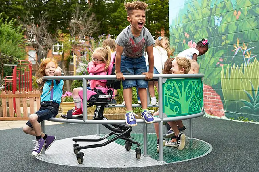 A group of children, including a wheelchair user, laughing and playing together on the green, wildlife-themed Inclusive Wheelspin Roundabout in a school playground.