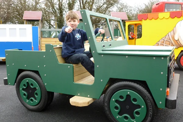 A side view of a green timber play jeep with a boy at the wheel, on a tarmac surface in an outdoor playground.
