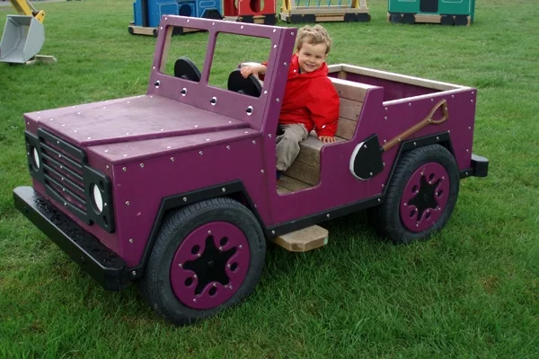 A side view of a purple timber play jeep with a boy inside, on a grassy area in an outdoor playground.