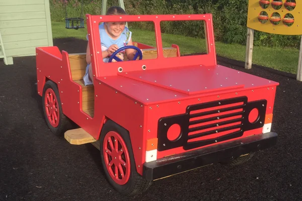 A front view of a red timber play jeep with a girl at the wheel, on a rubber surface in an outdoor playground.