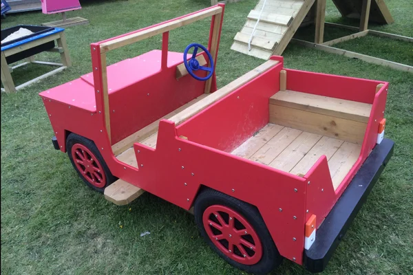 An aerial view of a red timber open-top play jeep with a blue steering wheel, on a grassy area in an outdoor playground.