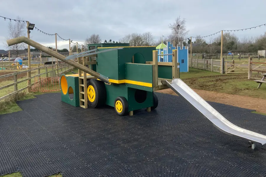 A green and yellow timber play combine harvester with tunnels and a slide, on a rubber surface in an outdoor playground.