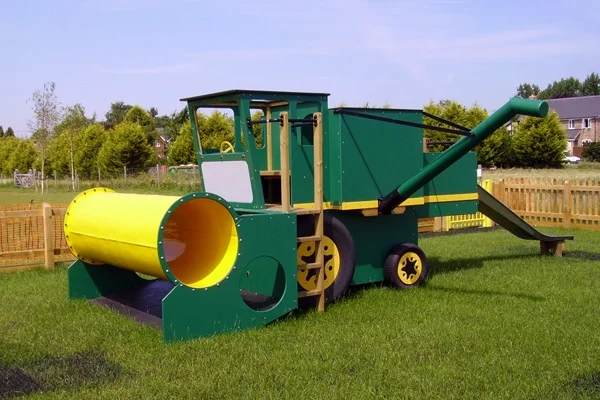 A green and yellow timber play combine harvester with a yellow tube, on a grassy area in an outdoor playground.