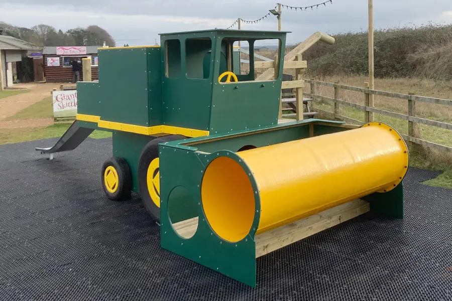 A front view of a green and yellow timber play combine harvester with a yellow tube, on a rubber surface in an outdoor playground.