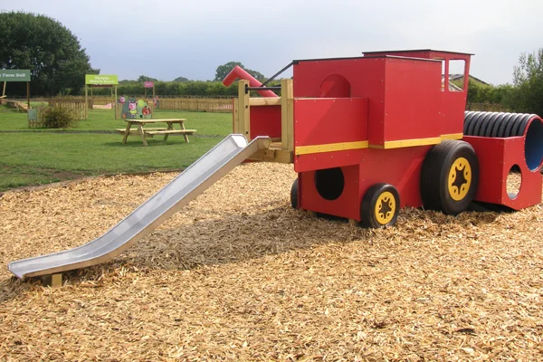 A red and yellow timber play combine harvester with a black tube slide, in a woodchip outdoor playground.