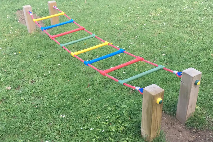 A colourful rope Ladder Traverse outdoor play equipment, with multi-coloured rungs, installed over a grassy area.
