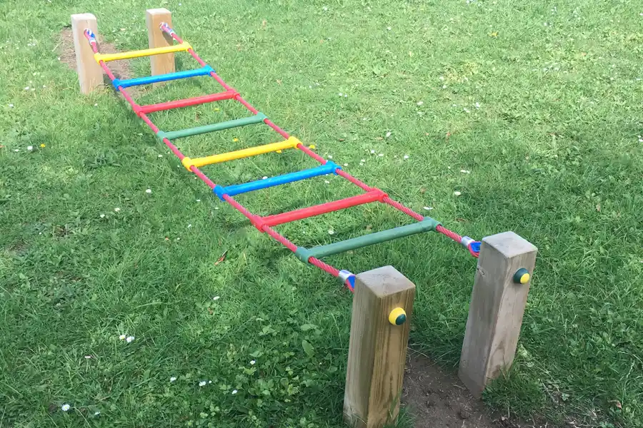A side view of the colourful rope Ladder Traverse, showing its low height and installation between two wooden posts in a grassy field.