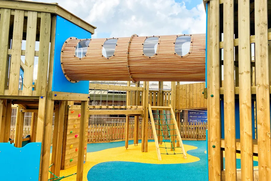 An elevated timber tunnel with porthole windows connecting two towers of the Ladram Towers multi-play unit, with blue panels, a rock climbing wall, and a rope net ladder visible below, on a yellow and blue rubber safety surface.