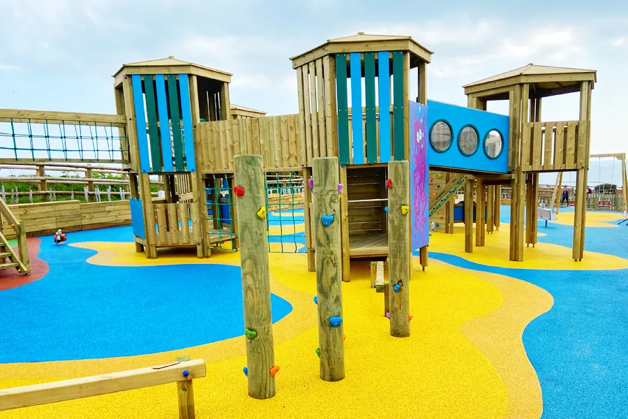 A wide view of the Ladram Towers multi-play unit showing three timber towers with teal and blue panels, timber climbing poles with coloured holds, a rope net, and a blue tunnel connector, on a yellow and blue rubber safety surface.