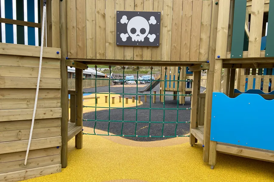 A pirate skull and crossbones panel above a rope net walkway connecting two towers of the Ladram Towers multi-play unit, with a rope climb, blue panels, and a yellow rubber safety surface, at a holiday park.