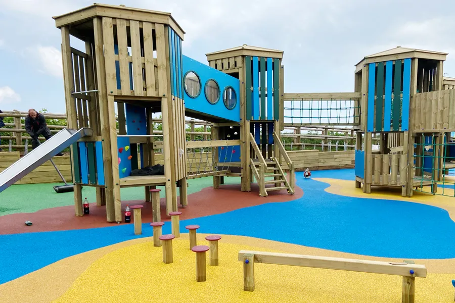 The Ladram Towers multi-play unit showing three timber towers with teal and blue panels, a stainless steel slide, a chain bridge walkway, a rock climbing wall, stepping posts, and a balance beam, on a yellow, blue, and red rubber safety surface.