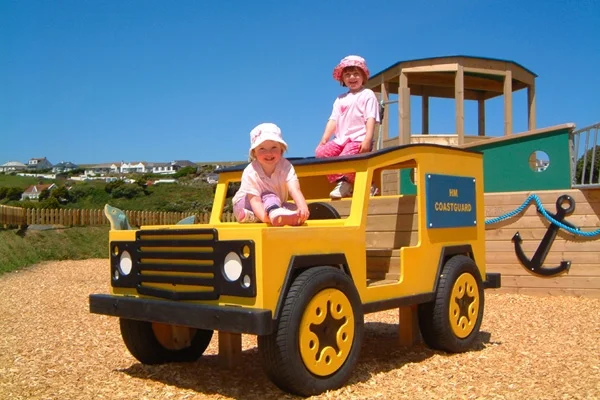 A front view of a yellow timber play Landrover with "HM Coastguard" livery, with two girls inside, in a woodchip outdoor playground.
