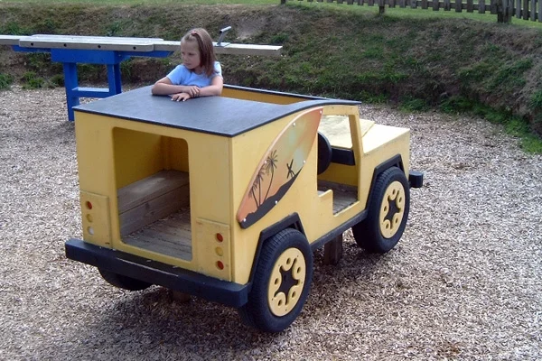 A rear view of a yellow timber safari-style play Landrover with a girl looking over the roof, in a woodchip outdoor playground.