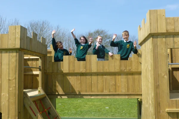 Four primary school children in green uniforms waving and smiling from the battlements of a Large Fort wooden castle play structure, on a sunny school playground with blue sky.