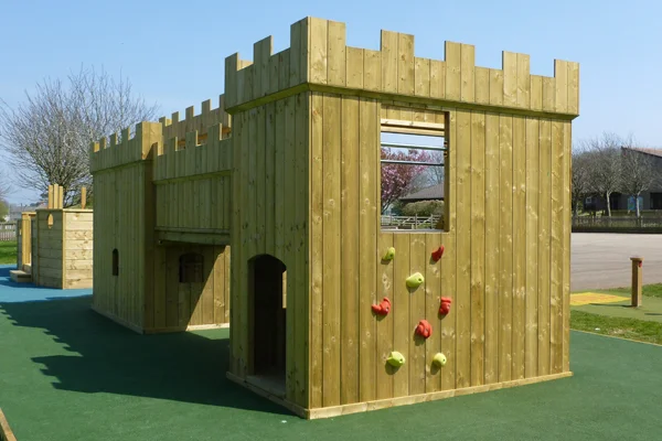 A close-up of the Large Fort wooden castle play structure showing the castle battlements, a colourful rock climbing wall with red and yellow holds, and a large arched doorway, on a green rubber safety surface.