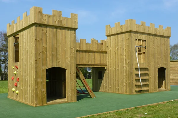 A full view of the Large Fort wooden castle play structure showing twin towers with battlements, a colourful rock climbing wall, a rope ladder, a drawbridge ramp, and arched doorways, on a green rubber safety surface.