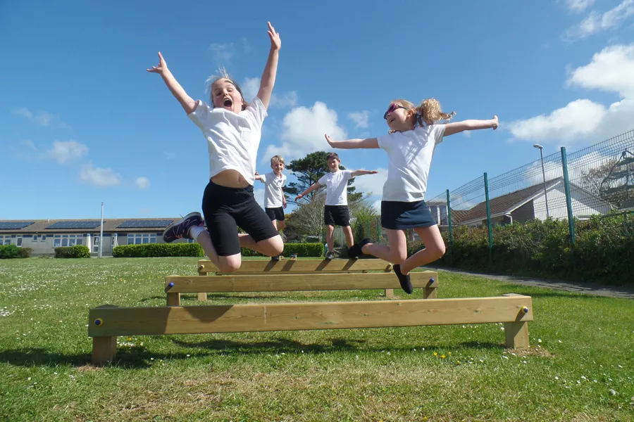 Two happy girls in school uniform jumping joyfully in mid-air over wooden Leaping Bars on a sunny school field.