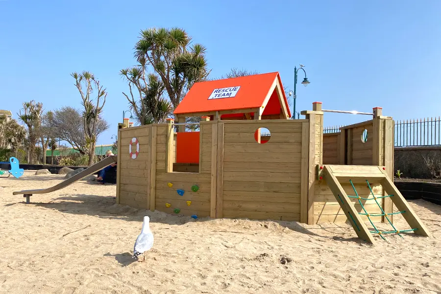 A seagull walks past the Lifeboat children's outdoor play structure on a sandy beach, showing the slide and climbing net.