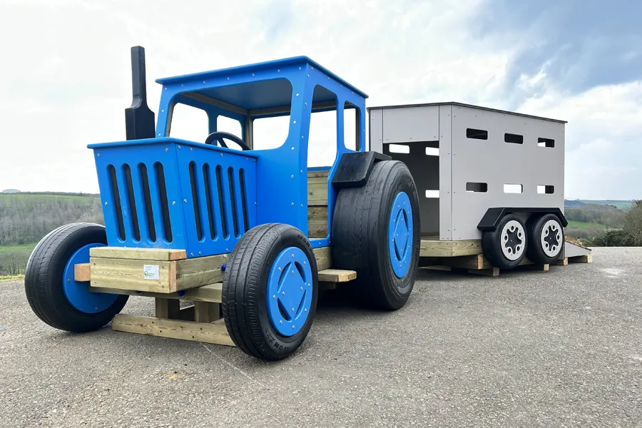 A grey timber play livestock trailer attached to a blue Tommy Tractor, on a tarmac surface in an outdoor playground.