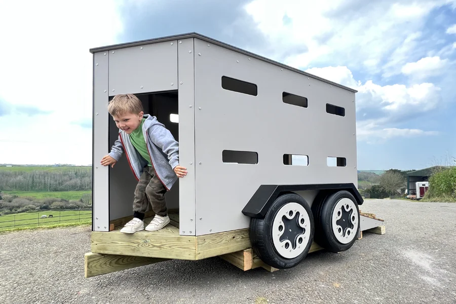 A young child climbing out of a grey timber play livestock trailer, on a tarmac surface in an outdoor playground.