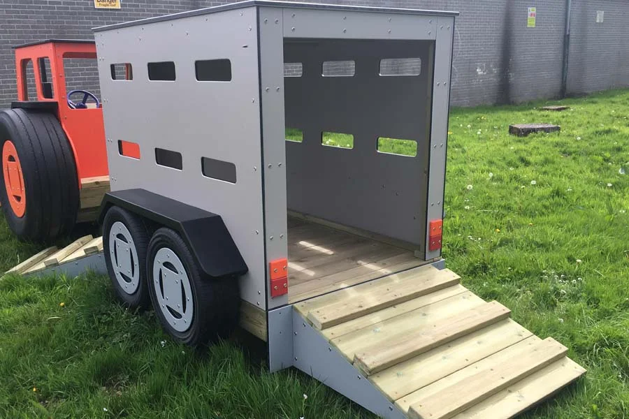 A grey timber play livestock trailer with an open door and timber loading ramp, on a grassy area in an outdoor playground.