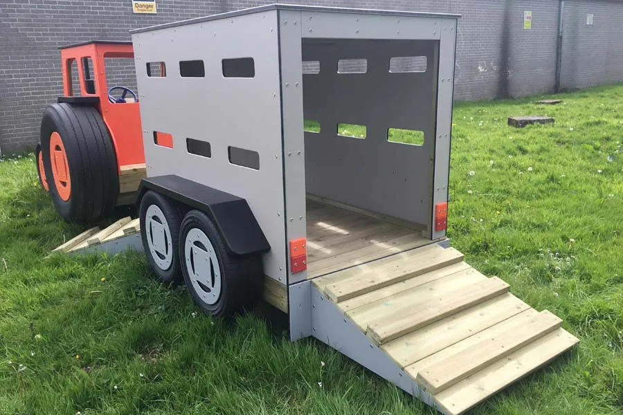 A grey timber play livestock trailer with an open door and timber steps, on a grassy area in an outdoor playground.