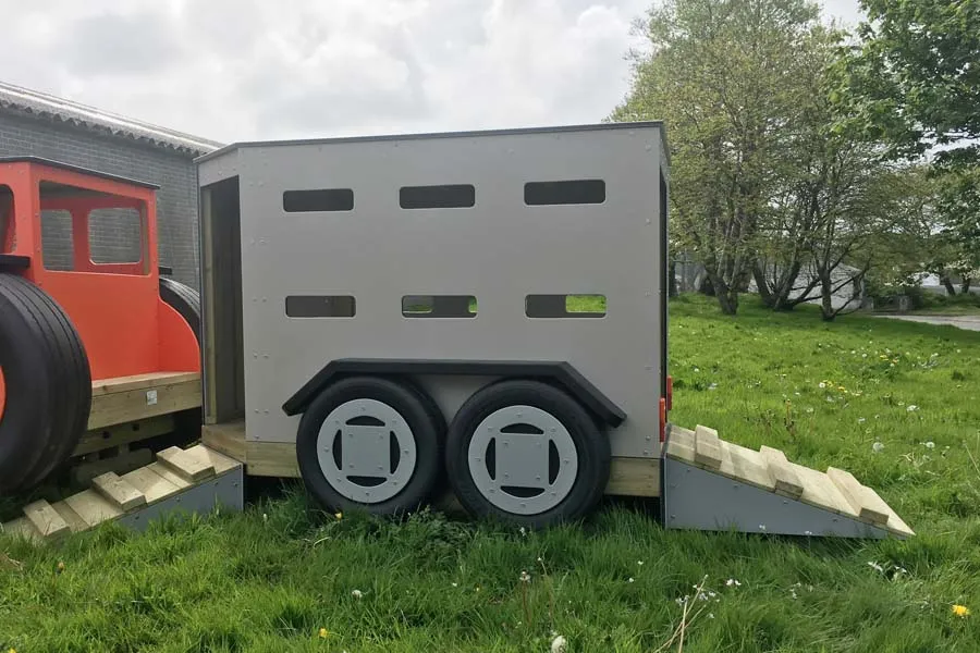 A rear view of a grey timber play livestock trailer with a loading ramp, next to a red Tommy Tractor on a grassy outdoor playground.