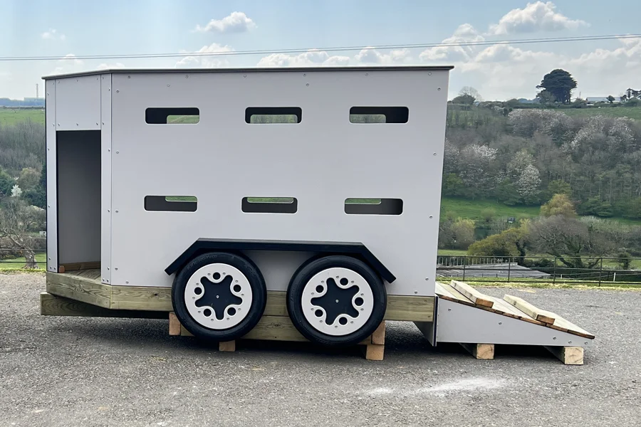 A white timber play livestock trailer with a loading ramp, on a tarmac surface in an outdoor playground.