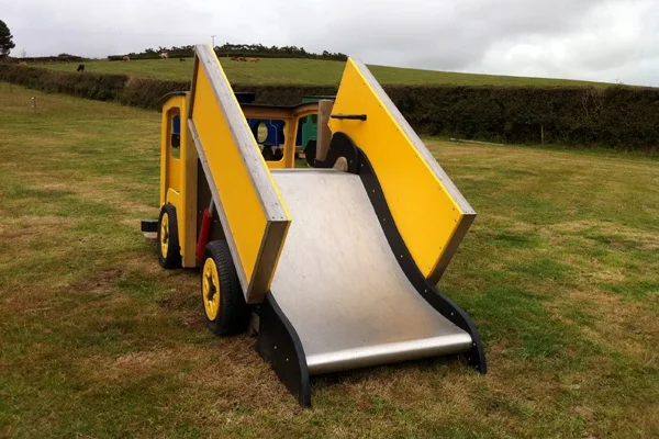 A front view of a yellow and black timber play lorry with a tipper truck slide, on a grassy area in an outdoor playground.