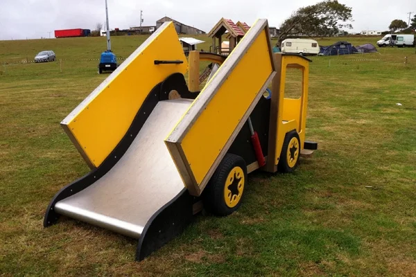 A rear view of a yellow and black timber play lorry with a tipper truck slide, on a grassy area in an outdoor playground.