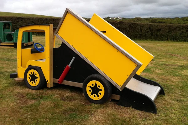 A side view of a yellow and black timber play lorry with a tipper truck slide, on a grassy area in an outdoor playground.