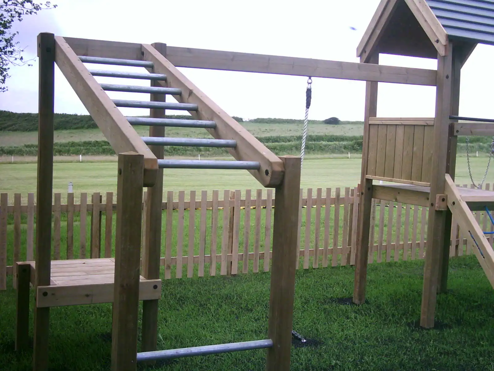 A close-up of the Maribou Play Frame showing the angled galvanised steel ladder, overhead monkey bars, and hanging rope swing, with a grey-roofed tower and coastal grassland visible in the background.