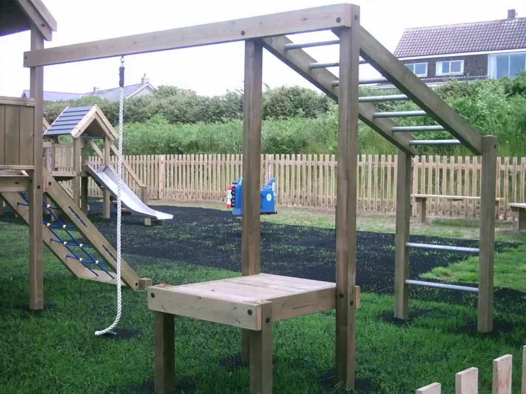 A close-up of the Maribou Play Frame showing the angled galvanised steel ladder, overhead monkey bars, and hanging rope swing, with a grey-roofed tower and coastal grassland visible in the background.