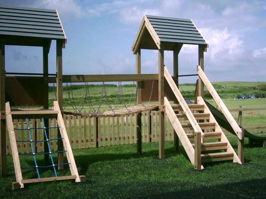 This side-angle view of the Maribou Play Frame shows the structure installed in a school playground surrounded by mature trees. The twin towers with distinctive red roofs, rope net climbing ramp with blue rungs, wooden staircase, chain bridge walkway, and galvanised monkey bars are all clearly visible, providing a comprehensive view of the structure's construction and play challenges.
