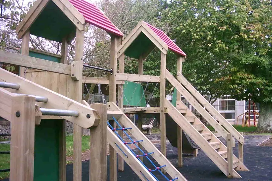 A Maribou Play Frame with twin towers with red roofs, a rope net climbing ramp, a wooden staircase with blue rungs, a chain bridge walkway, and galvanised monkey bars, installed in a school playground with trees.