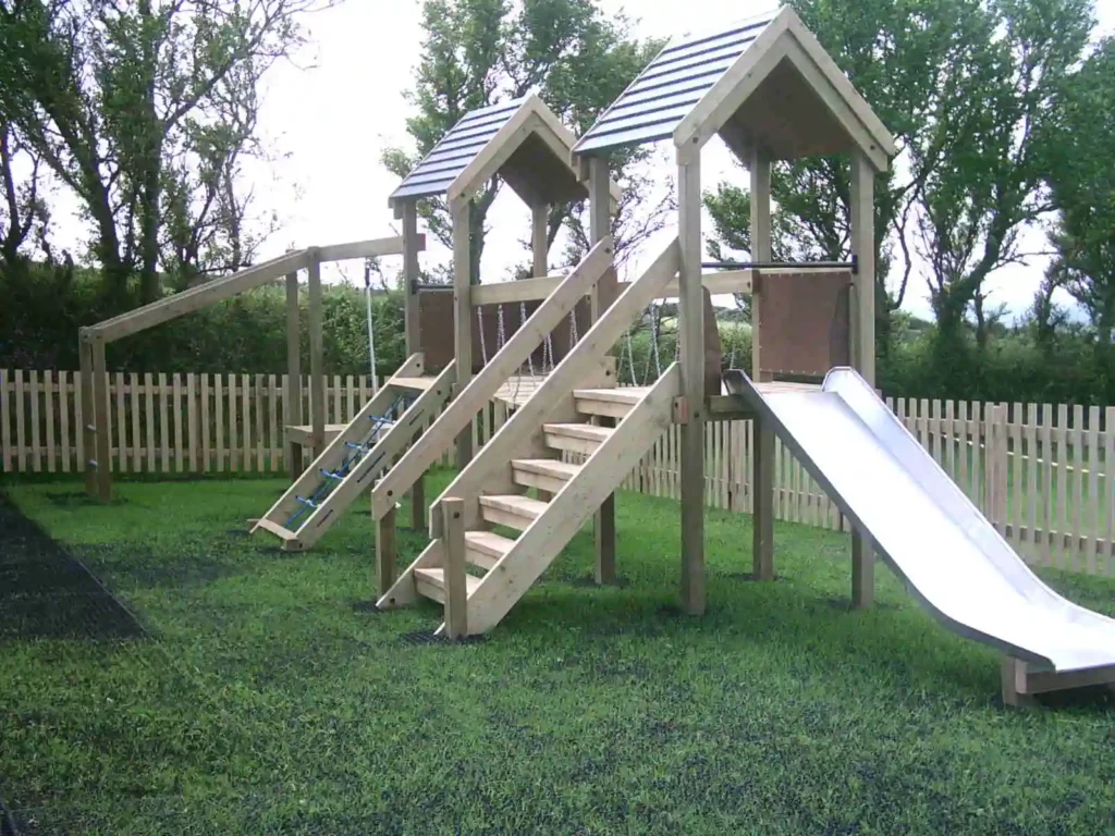 A Maribou Play Frame with twin towers with grey roofs, a wooden staircase, a stainless steel slide, a rope net climbing ramp, and a chain bridge walkway, installed on a grass playground with a picket fence and trees.