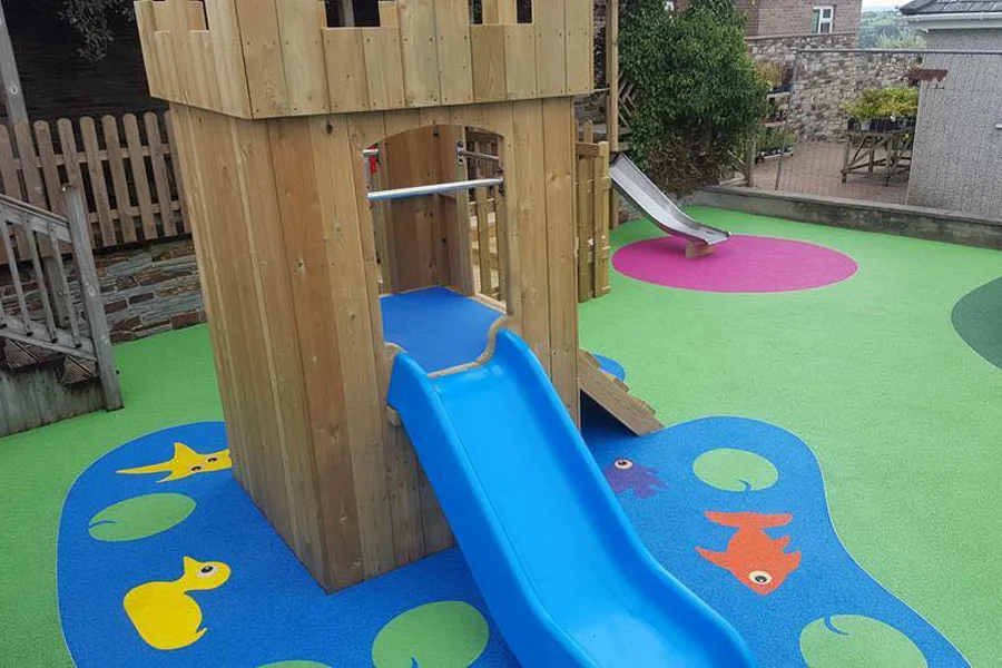 A wooden Mini Fort castle play unit with battlements and an arched doorway, featuring a blue plastic slide, on a blue rubber safety surface with colourful fish motifs, in an outdoor playground with stone walls.