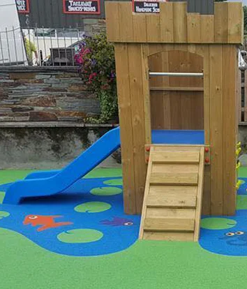 A wooden Mini Fort castle play unit with battlements and an arched doorway, featuring a blue plastic slide, on a blue rubber safety surface with colourful fish motifs, in an outdoor playground with stone walls.
